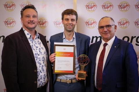 Three men standing in front of EMPA banner. Middle man, Nathan Maddock holding an EMPA award