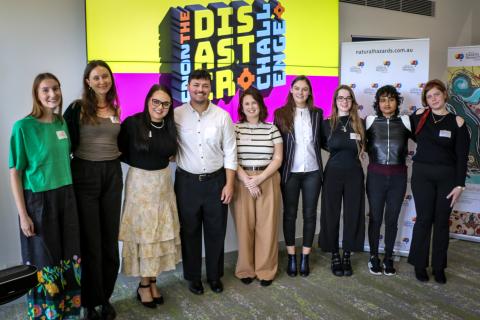 9 people standing in a row smiling in front of large TV screen with Disaster Challenge logo